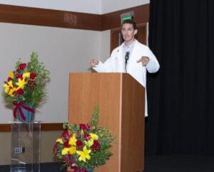 A person in a white coat speaks at a podium on a stage, with two large floral arrangements nearby.