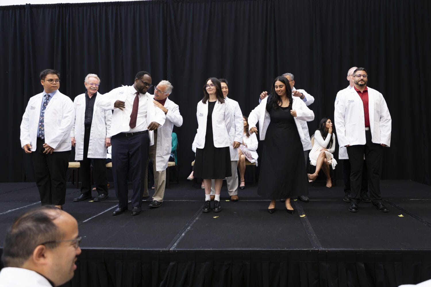 A group of people on a stage put on white coats during a ceremony, with others seated in the background against black curtains.