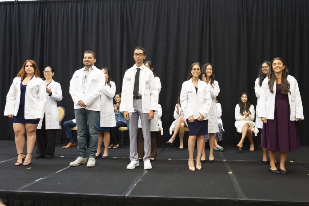 A group of students in white coats stand on a stage, smiling, with others seated behind them against a black curtain backdrop.