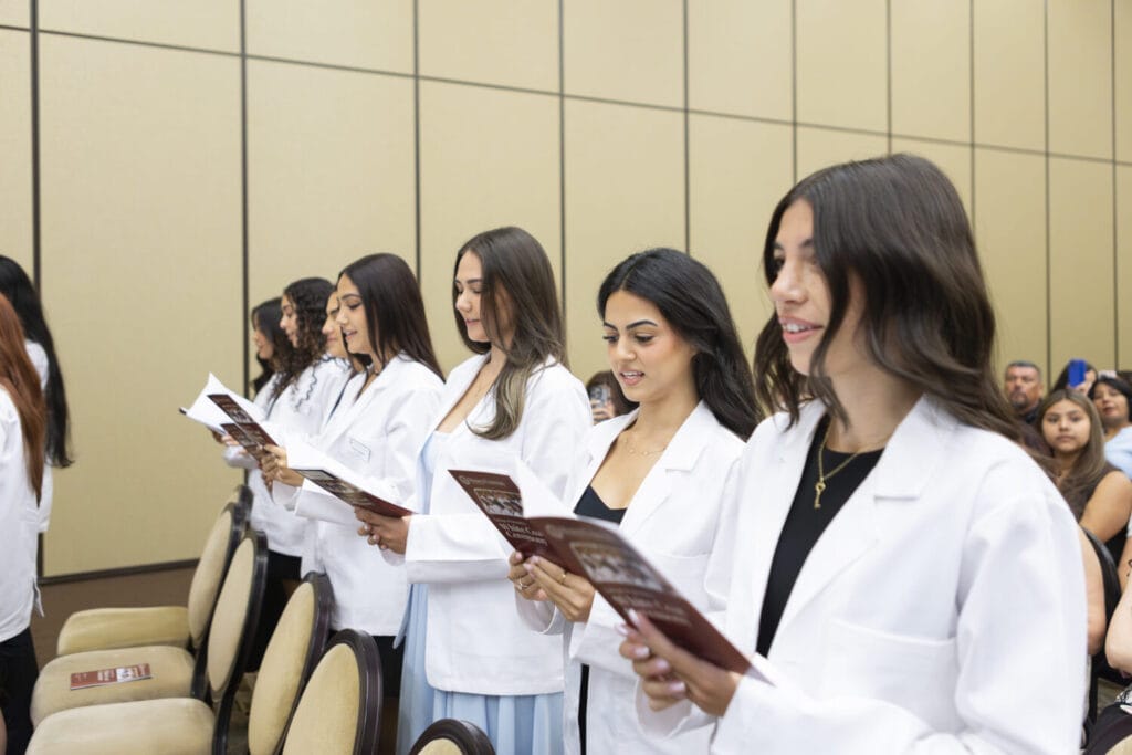 A group of students in white coats stands in a row, holding booklets and reading during a ceremony in a conference room.
