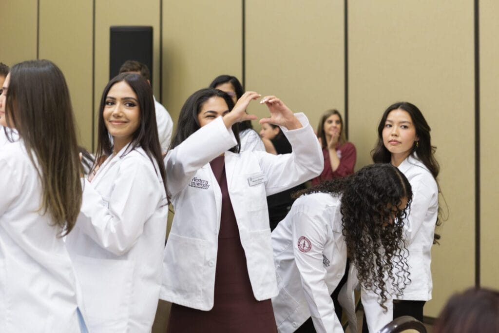 A group of people wearing white lab coats stand indoors; one person in the center makes a heart shape with their hands.