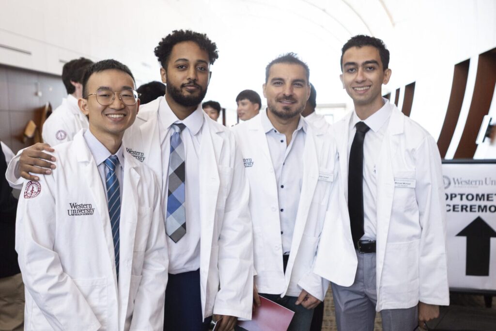 Four men wearing white lab coats and formal attire stand together, smiling at the camera in an indoor setting with a sign reading "Optome..." visible in the background.