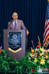 A man in a suit speaks at a podium with a Western University College of Podiatric Medicine banner, surrounded by flowers and a U.S. flag.