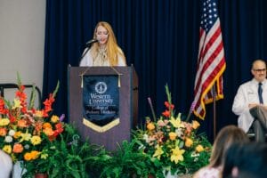 A woman speaks at a podium labeled "Western University College of Podiatric Medicine," with floral arrangements and the American flag nearby; a man sits to the right.
