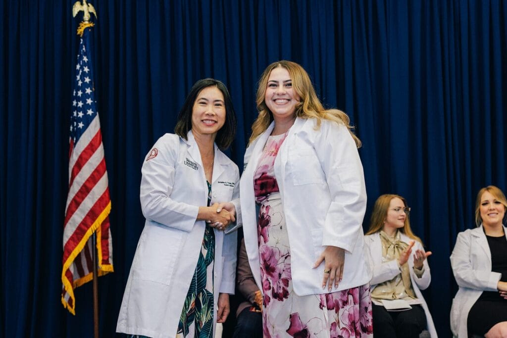 Two women in white coats shake hands in front of a blue curtain and an American flag; people in white coats are seated and clapping in the background.