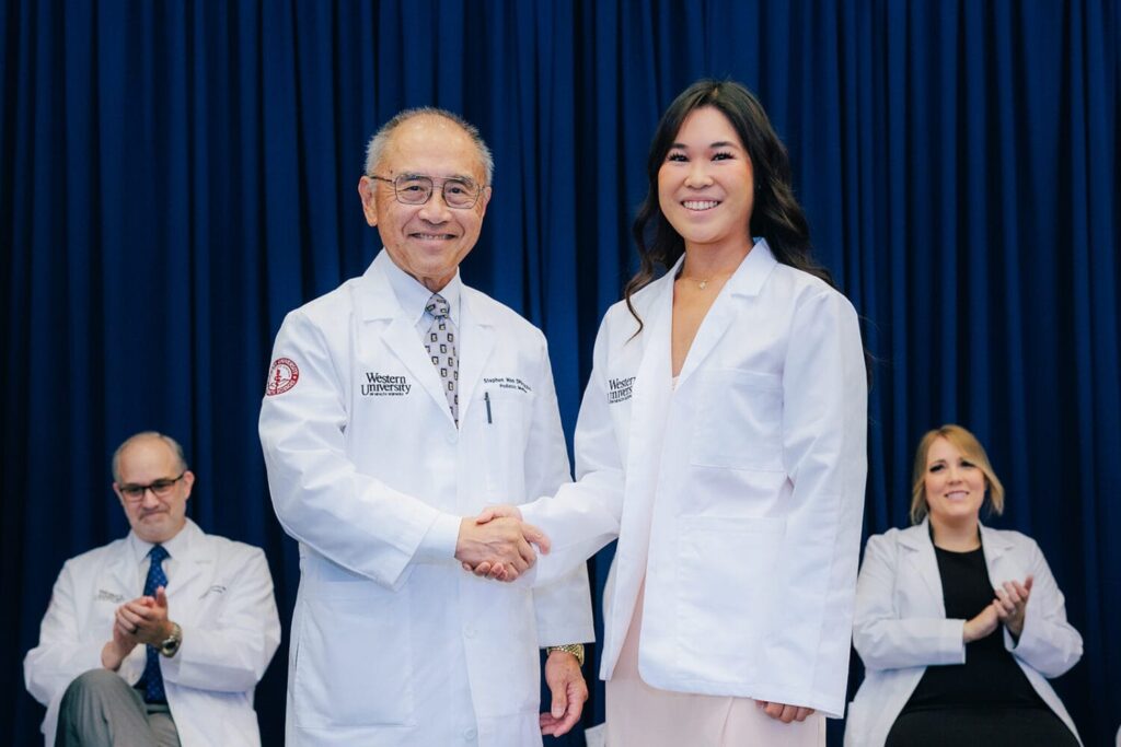 An older man in a white coat shakes hands with a young woman in a white coat on stage, while two seated people applaud in the background.