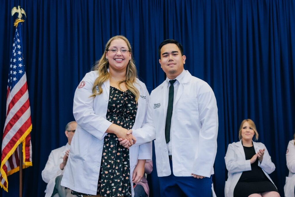 Two people in white coats shake hands on stage in front of an American flag and blue curtain, with others seated and applauding in the background.