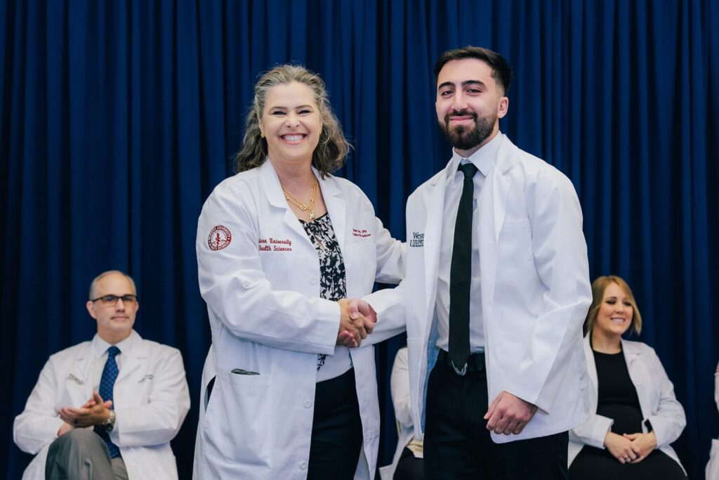 Two people in white coats shake hands and smile on stage, with two seated individuals in the background against a blue curtain.