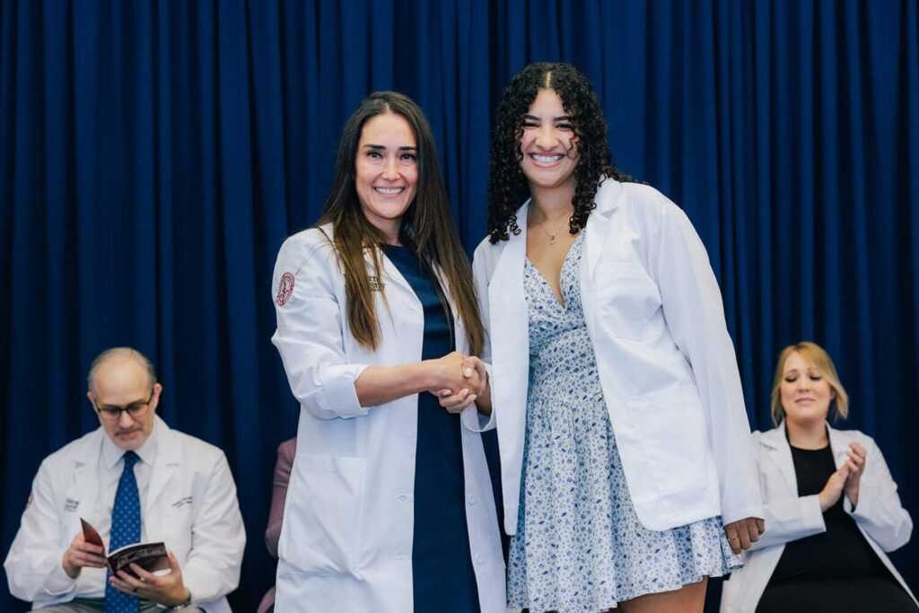 Two women in white coats pose and shake hands on stage, smiling at the camera, with two seated people in white coats in the background.