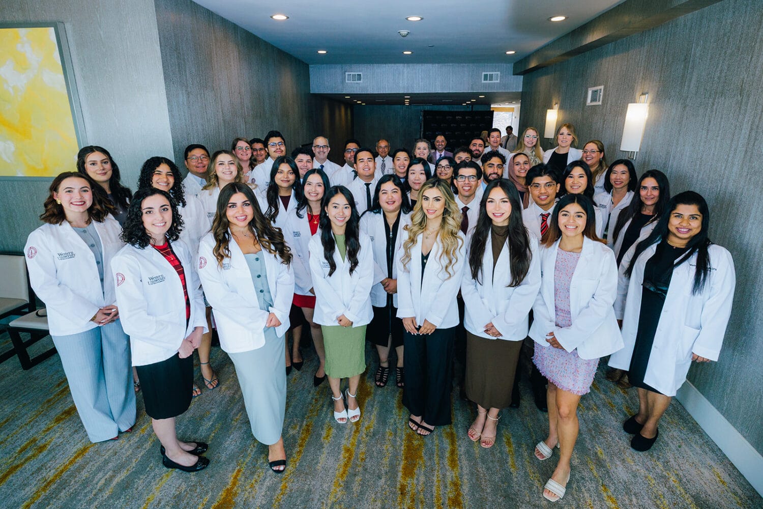 A large group of people, many wearing white coats, pose together in a hallway, likely for a professional or academic event.
