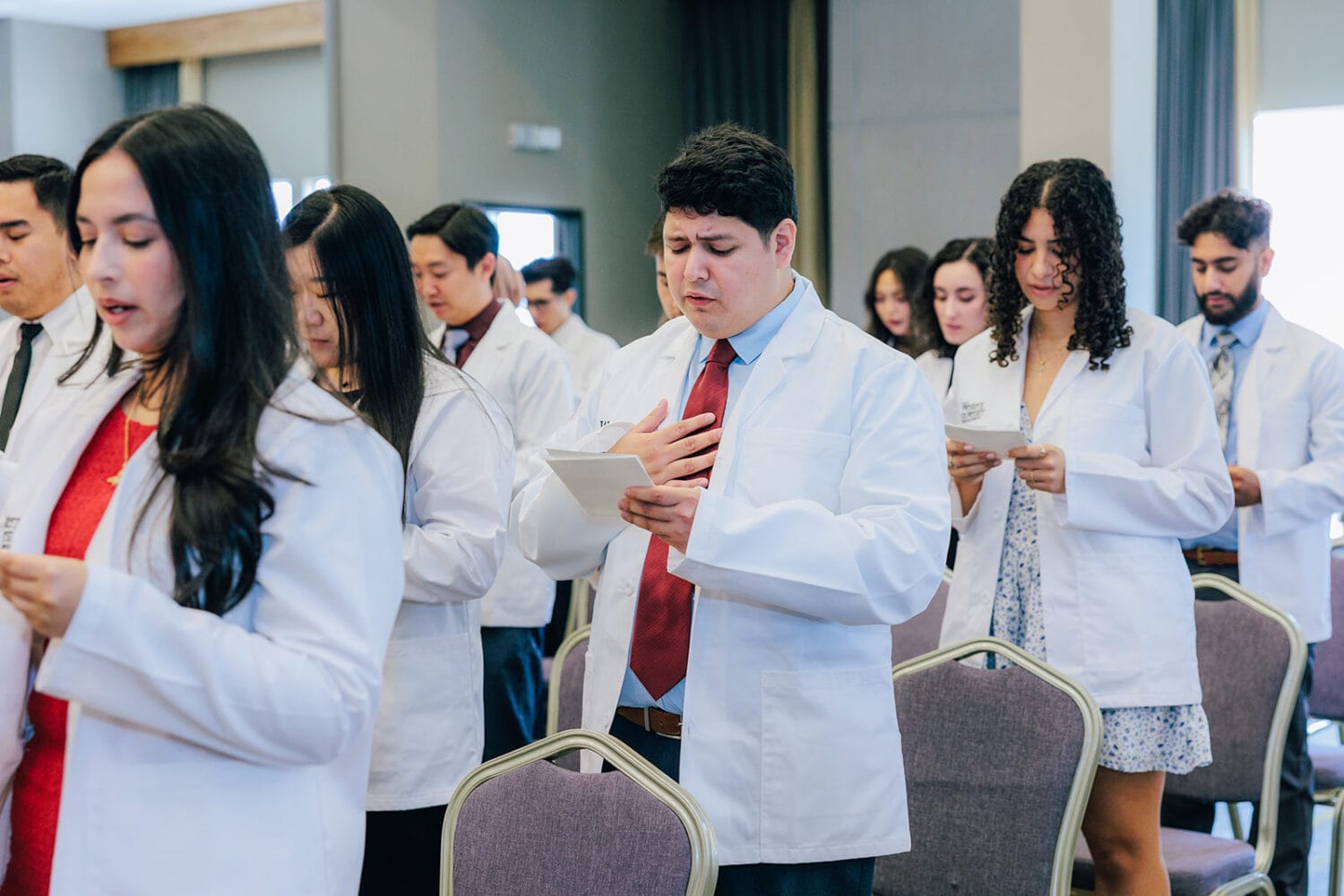A group of people in white coats stand and read from papers during a formal event, with one person holding a hand to their chest.