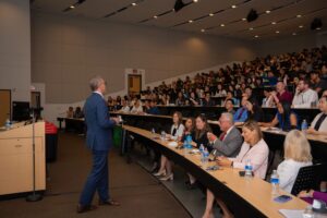 A speaker in a suit addresses a large audience seated in a tiered lecture hall. Attendees listen attentively, with notebooks and bottled water on desks.