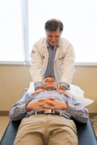 A doctor in a white coat performs a physical examination on a patient lying on an exam table with hands folded on his stomach.