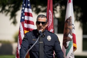 A police officer in uniform and sunglasses speaks at a podium outdoors, with the U.S., California, and a university flag in the background.
