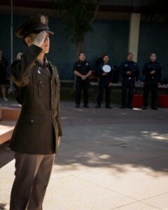 A uniformed officer salutes in the foreground while several people, some in police uniforms, stand in the background during an outdoor event.
