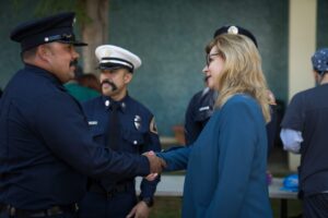 A woman in a teal blazer shakes hands with a uniformed police officer while other officers stand nearby.