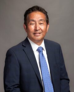 Man in a blue suit, white shirt, and patterned blue tie poses against a neutral gray background, looking at the camera with a slight smile.