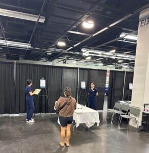 A woman stands in front of a table staffed by two healthcare workers in a room with black curtains and several chairs.