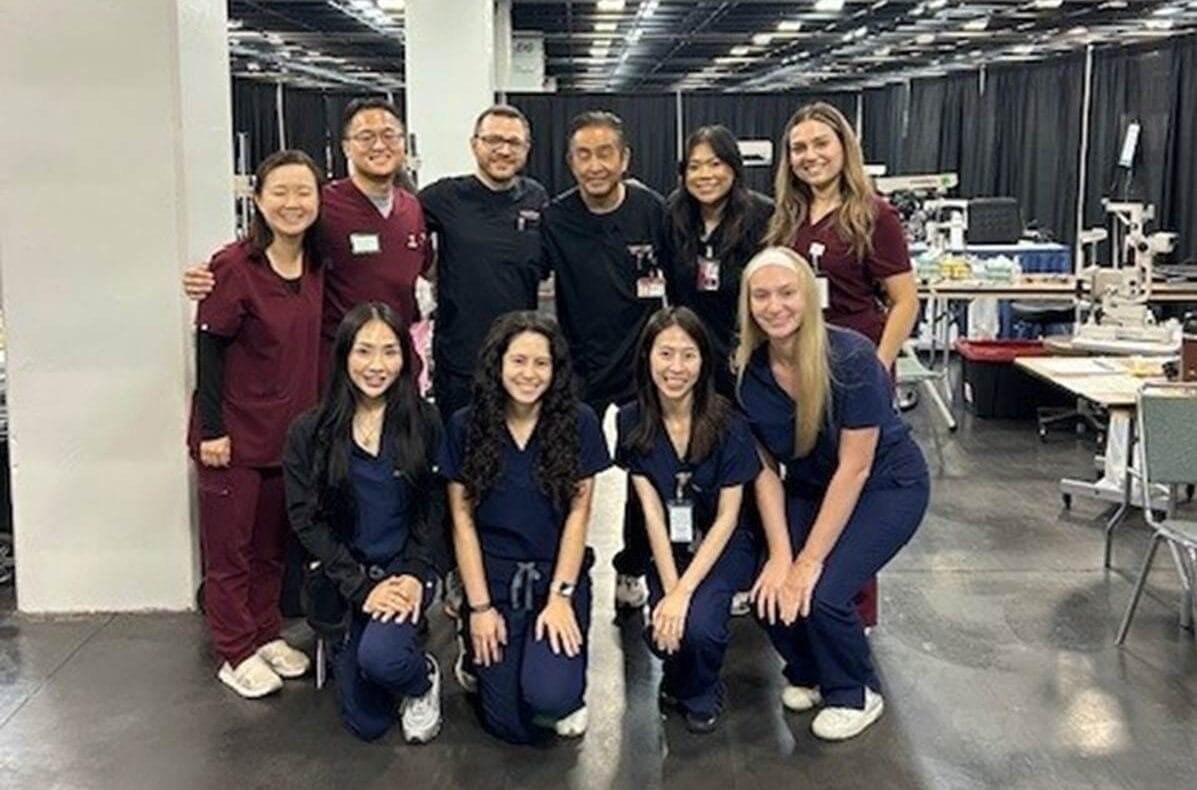 A group of healthcare workers in scrubs pose and smile together indoors at a convention center or clinic setting.