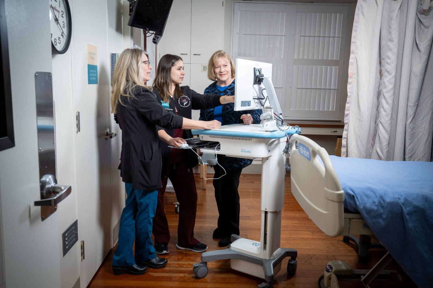 Three medical professionals stand beside a hospital bed, looking at a monitor on a mobile workstation in a clinical room.