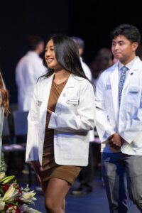 A woman in a white coat and brown dress smiles while standing next to a man in a white coat at a ceremony, with others in white coats in the background.