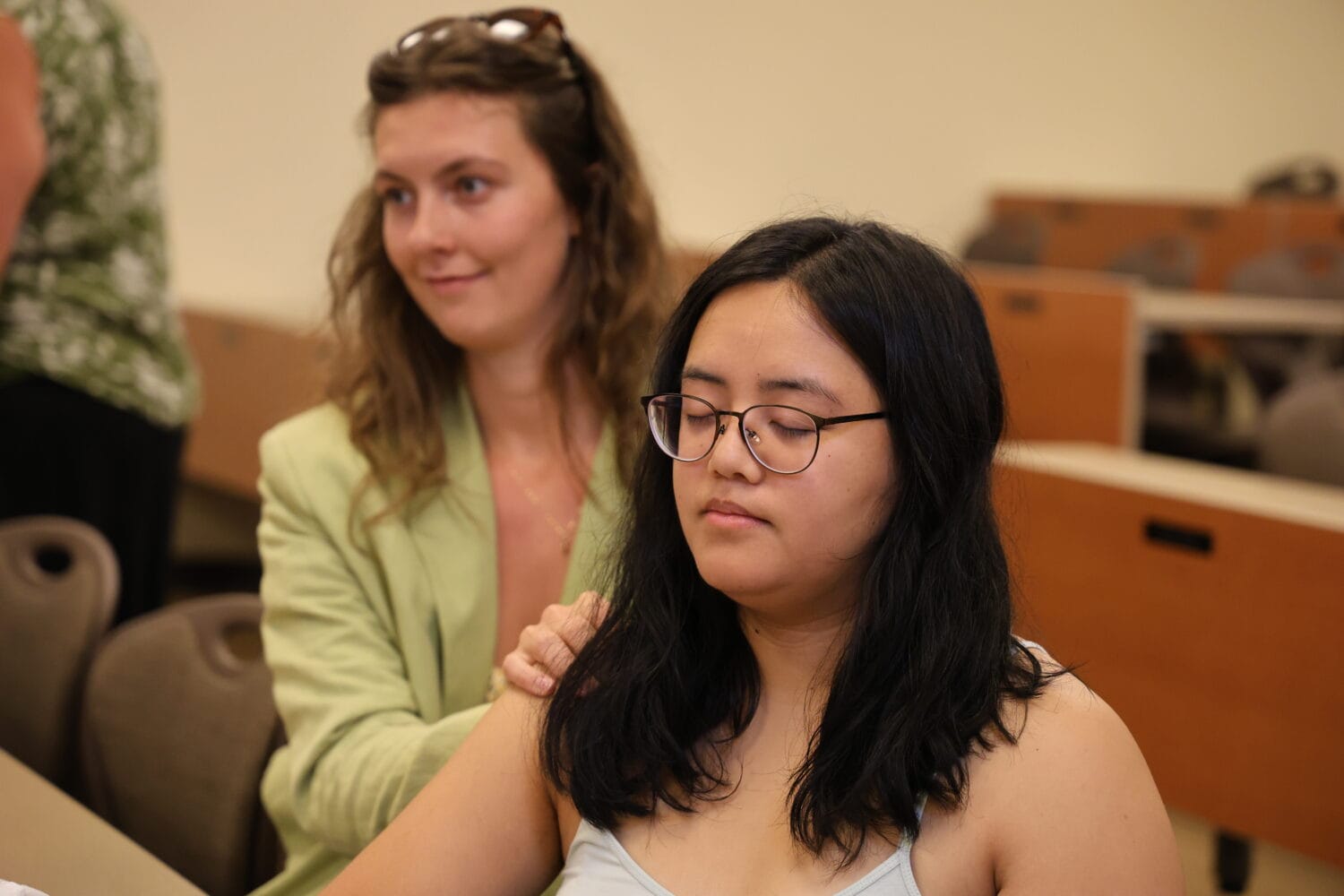 A woman places her hand on the shoulder of another woman with closed eyes, seated in a classroom with brown desks and chairs in the background.