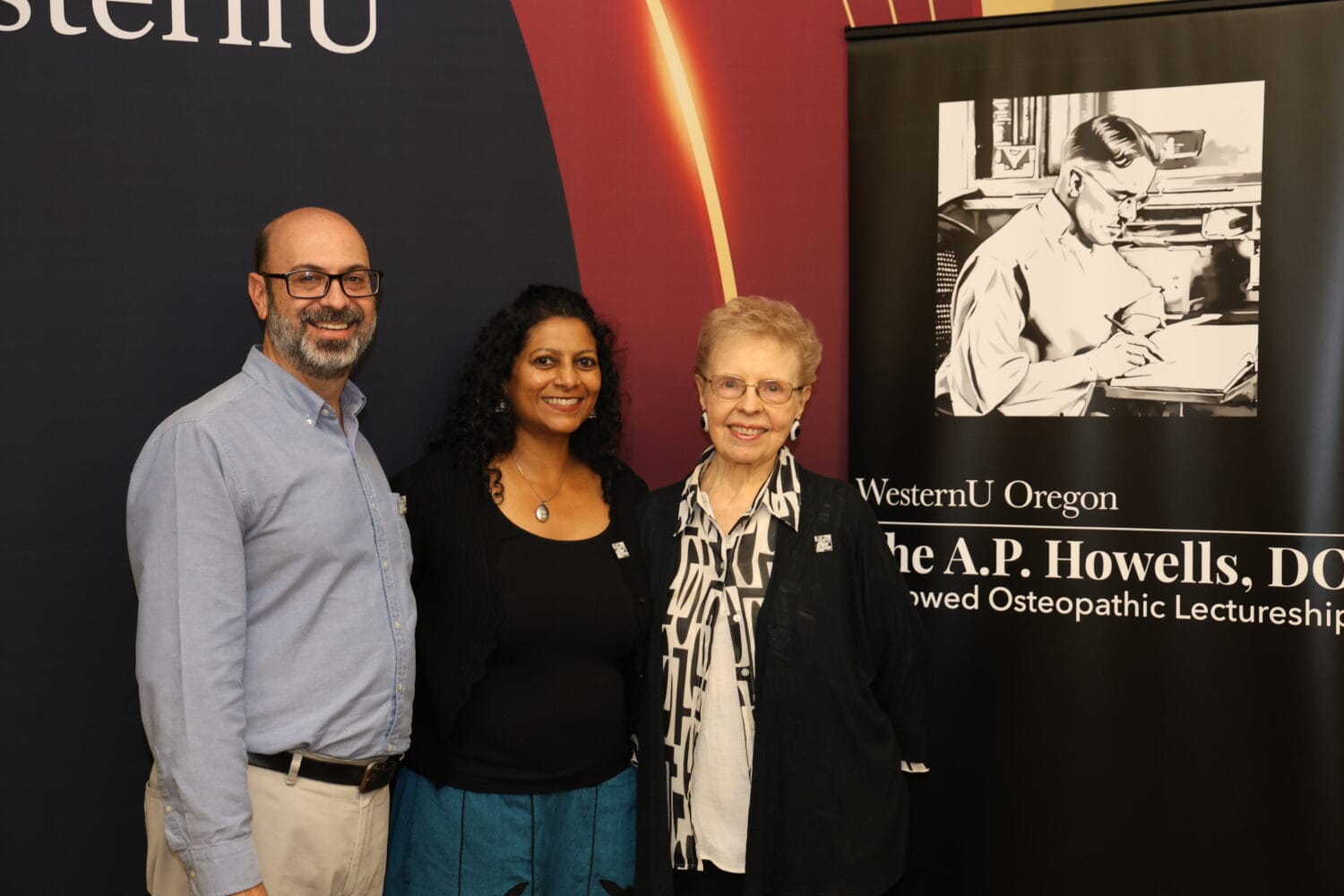 Three people stand side by side, smiling, in front of a WesternU banner and a poster featuring an illustration of A.P. Howells, DO.
