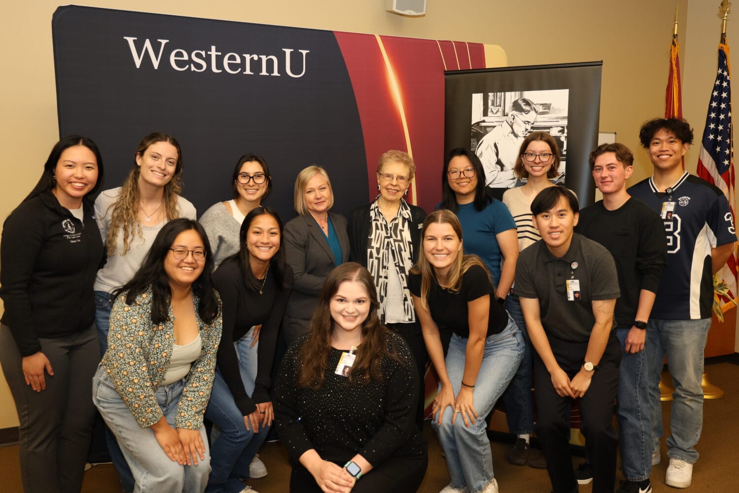 A group of people pose and smile for a photo in front of a WesternU banner in an indoor setting.