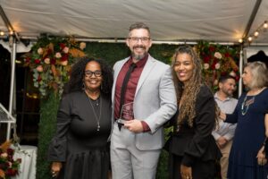 Three people pose together at an event; the man in the center is holding an award. A green floral backdrop and others are visible in the background.