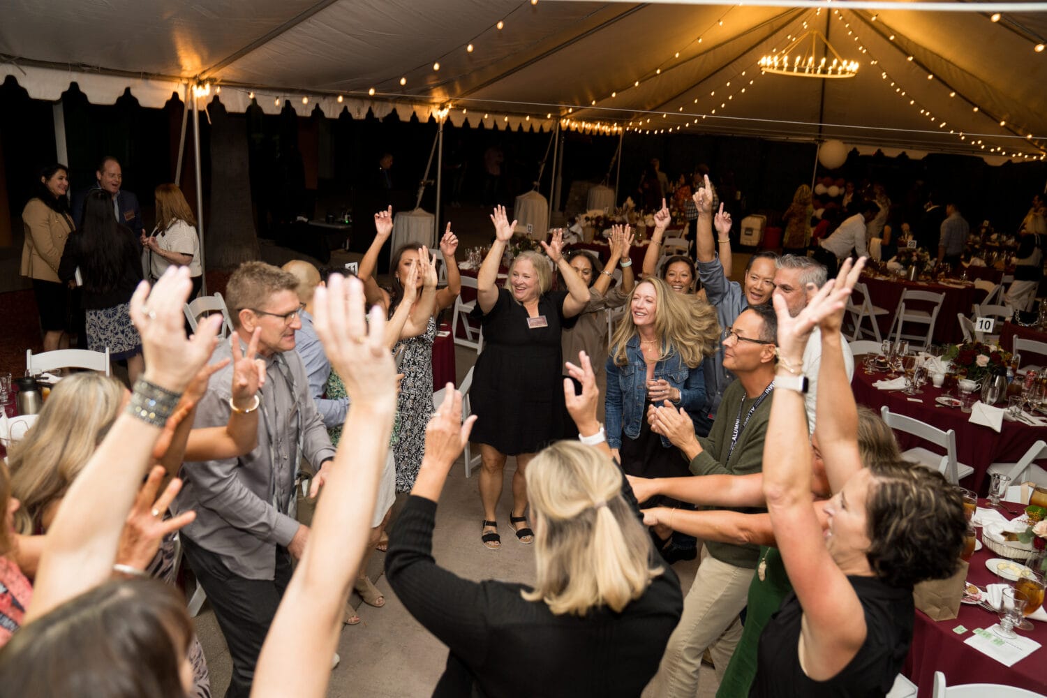 A group of people stand in a circle under a large event tent, raising their hands and smiling, with decorated tables and string lights in the background.