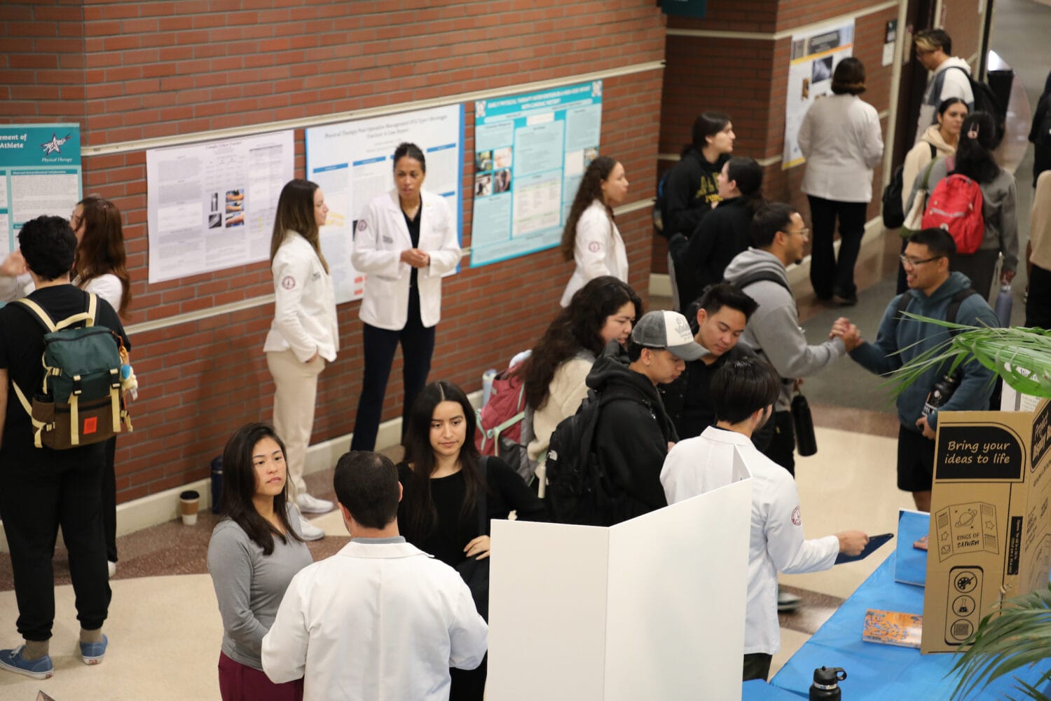 People in lab coats and visitors interact at an indoor science fair or academic event with posters and informational booths displayed along a brick wall.