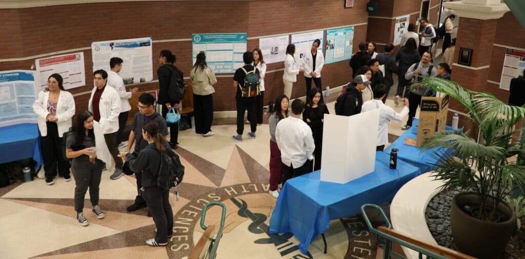 People gather in a university lobby with informational posters and tables set up for a science or health fair event. Some attendees are wearing white lab coats.