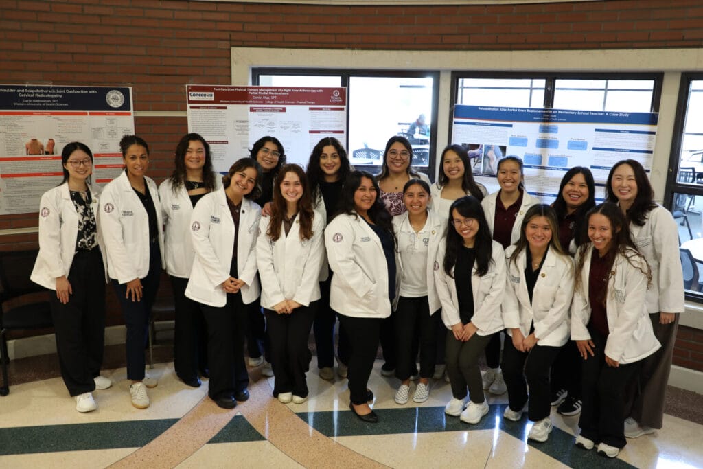 A group of students wearing white lab coats stand together and smile in front of research posters displayed on a brick wall indoors.