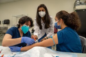 A healthcare worker draws blood from a seated patient’s arm while another medical professional observes; all three are wearing masks and gloves in a clinical setting.