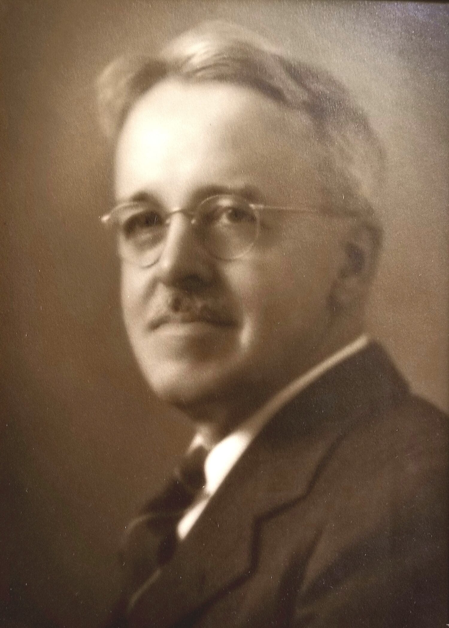A sepia-toned portrait of an older man with glasses, light hair, and a mustache, dressed in a suit and tie, looking slightly to the side.