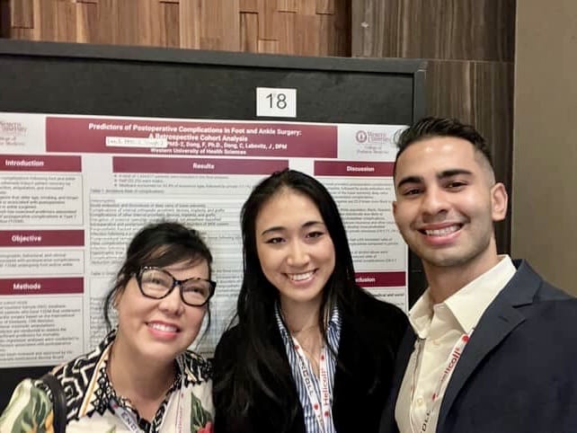 Three people stand in front of a scientific research poster titled "Predictors of Postoperative Complications in Foot and Ankle Surgery" at an academic conference.