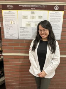 A woman in a white coat stands and smiles in front of a research poster about knee pain rehabilitation, displayed on a brick wall.