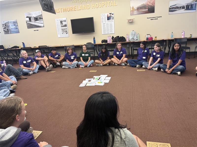 A group of students sit in a circle on a brown carpeted floor, facing papers and objects placed in the center of the circle in a classroom setting.