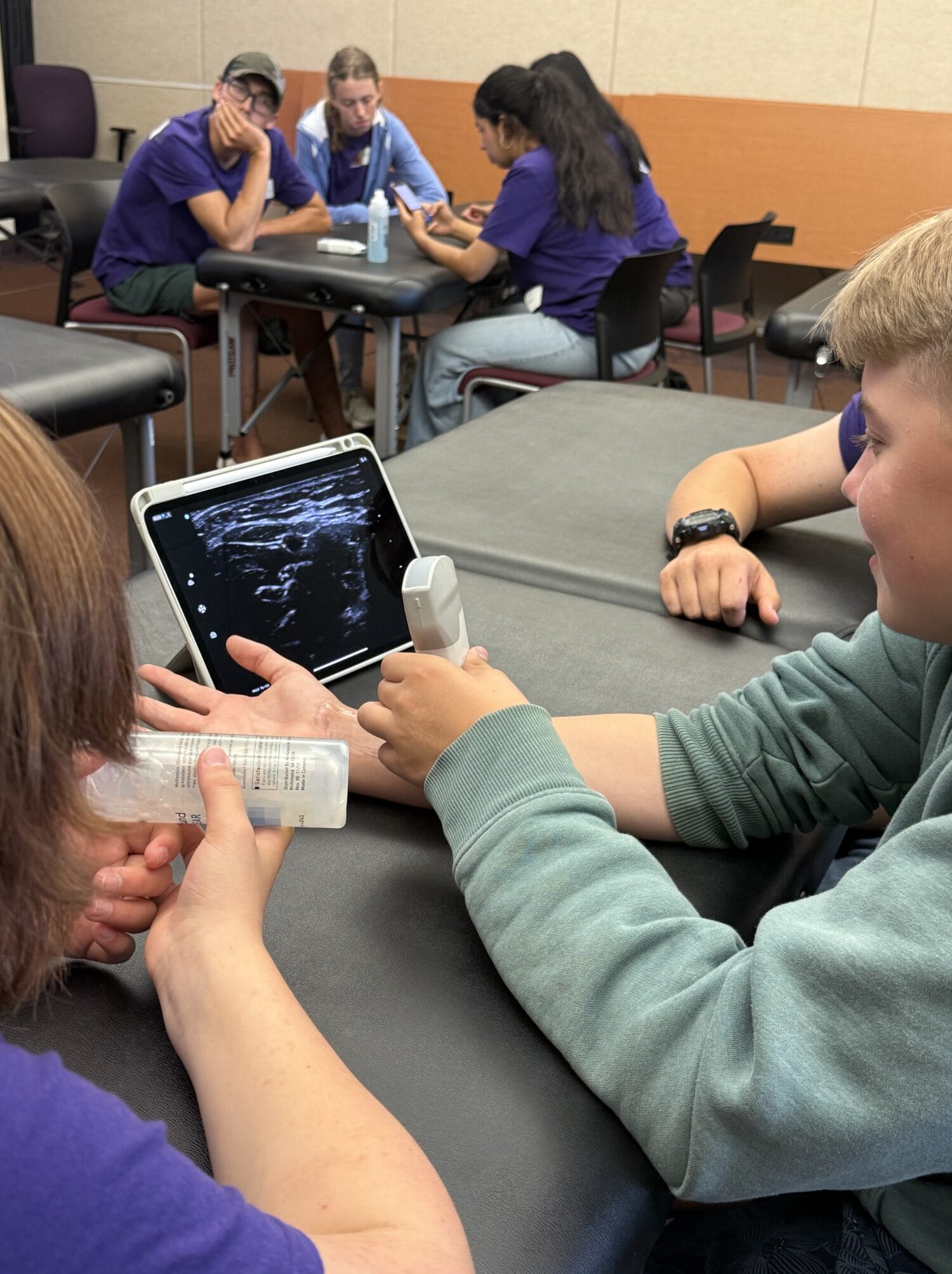 A student uses an ultrasound device on another person's arm while viewing the scan on a tablet in a classroom setting. Other students sit at tables in the background.