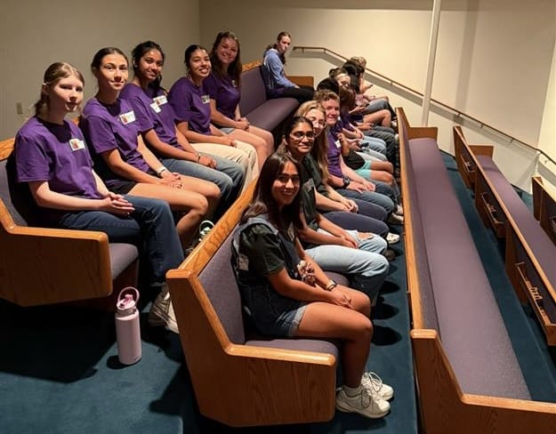 A group of young people sits in two rows of wooden benches in an auditorium, most wearing purple shirts, facing forward and smiling at the camera.