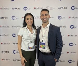 Two people dressed in business attire stand and smile in front of a step-and-repeat banner with multiple sponsor logos at an event.