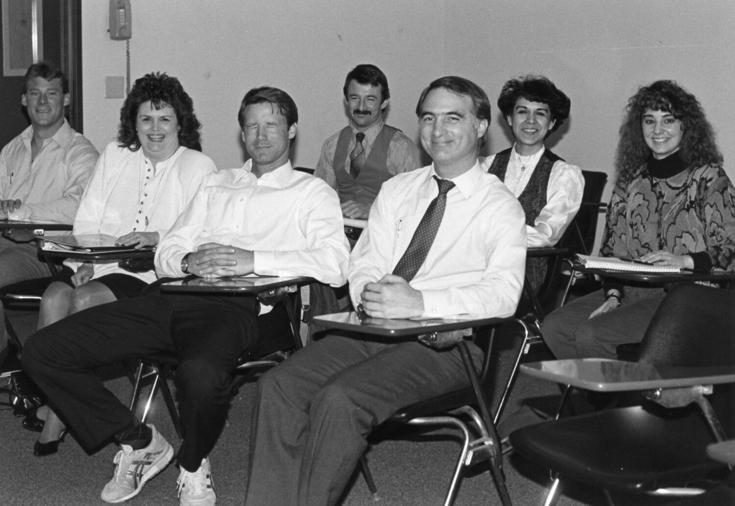 Seven adults sit in desks arranged in two rows in a classroom, facing the camera and smiling. The setting appears formal and academic.