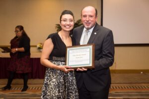 Two people standing indoors, smiling and holding an award plaque together. A person in the background appears to be preparing something at a table.