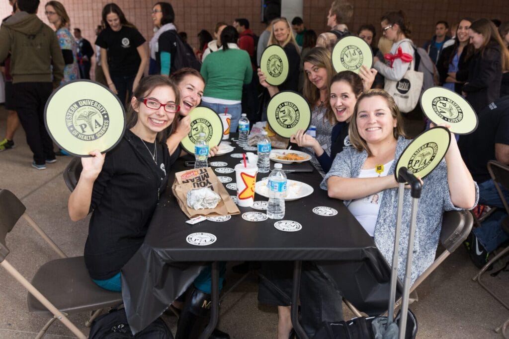 A group of people sit around a table at an event, holding circular university-branded signs and smiling at the camera. Food, drinks, and event materials are on the table.
