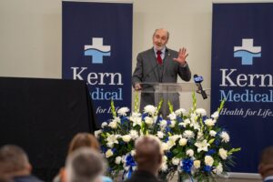 A man in a suit speaks at a podium with flower arrangements, in front of banners reading "Kern Medical" and "Health for Life." People are seated in the audience.