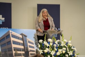 A woman stands at a podium speaking, with a floral arrangement and a poster of a medical building labeled "Kerr Medical" in front of her.