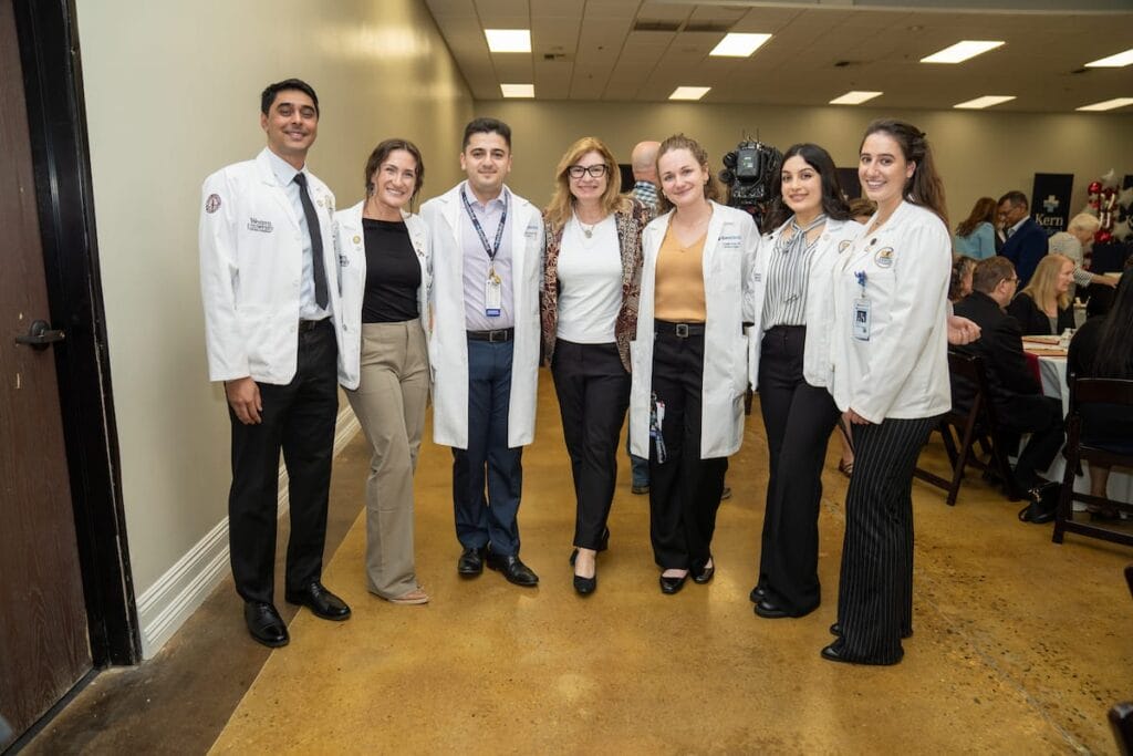 A group of seven people, including medical professionals in white coats and business attire, stand together and pose for a photo in a conference room.