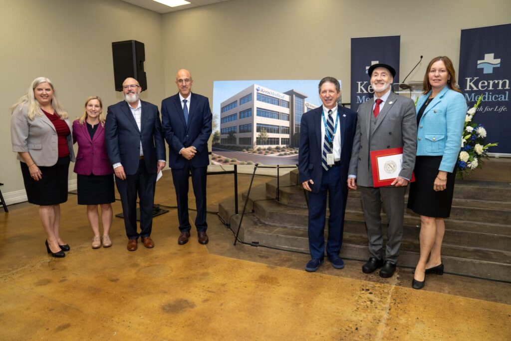 A group of seven people pose indoors next to a display board featuring an image of a Kern Medical building during an event.