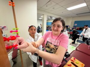 A woman in a pink shirt participates in a physical therapy exercise with a therapist’s assistance in a clinical setting.