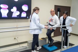 A man practices balance exercises on a blue foam pad between parallel bars, assisted by two healthcare professionals in a clinic setting.